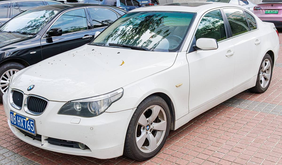 Bílý sedan BMW řady E60 zaparkovaný na dlážděném parkovišti, pohled zepředu z boku
