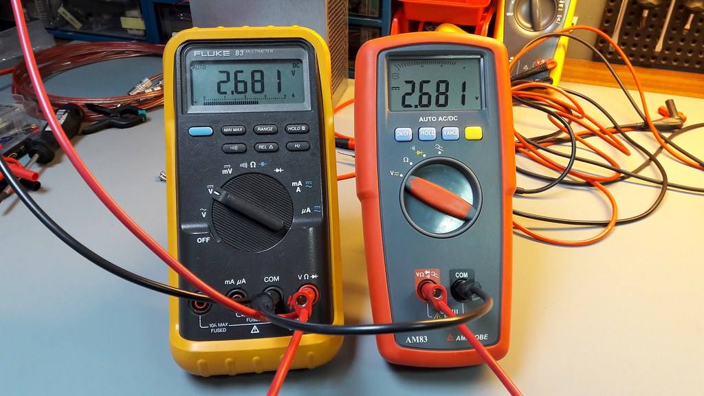 Dva digitální multimetry, žlutý Fluke 83 a oranžový Amprobe AM83, na dílenském stole s připojenými měřicími kabely