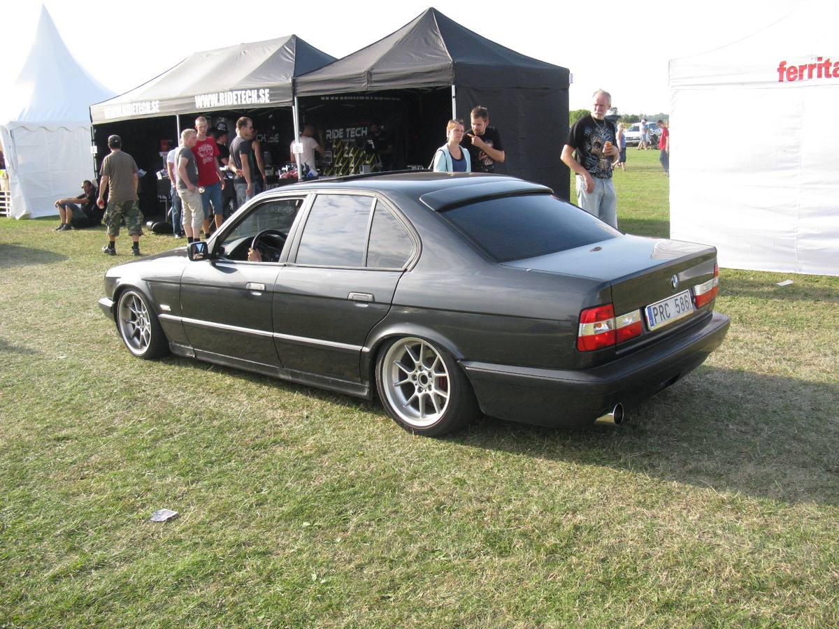 Tmavě zelené BMW 525i řady E34 zezadu ze strany, vystavené na trávě u stanů