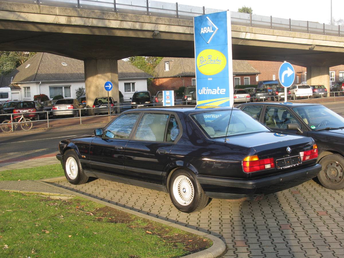 Tmavě modré BMW 750iL řady E32 zezadu ze strany, zaparkované u čerpací stanice pod mostem