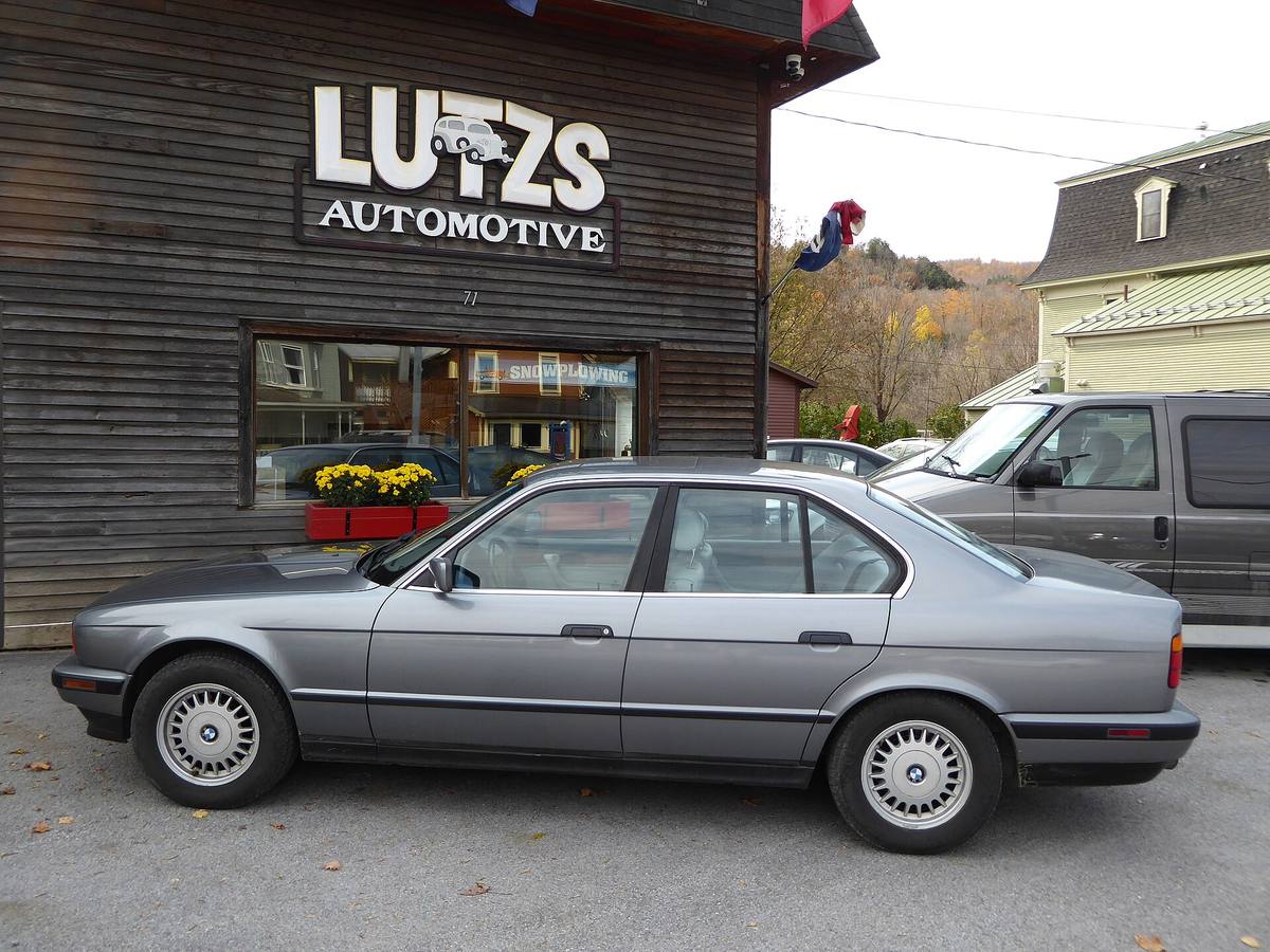 Šedý sedan BMW 525i řady E34 z boku, zaparkovaný před dřevěnou budovou autoservisu