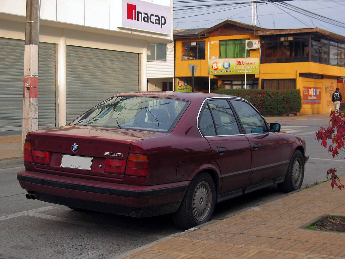 Vínově červený sedan BMW 530i z řady E34 zaparkovaný u chodníku, pohled zezadu zboku