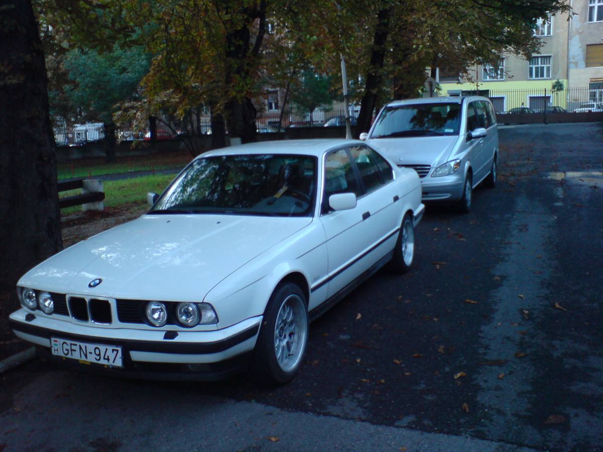 Bílý sedan BMW 525i řady E34 zaparkovaný pod stromy